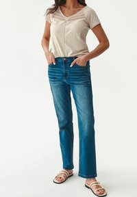 Top de manga corta beige con detalles de encaje, combinado con jeans rectos azules. El modelo lleva sandalias blancas con tiras.