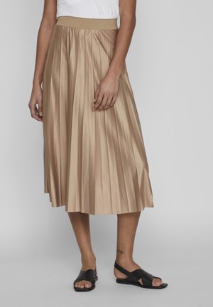 Femme portant une jupe midi plissée beige, un haut blanc et des sandales noires à bout ouvert, debout devant un fond gris uni.