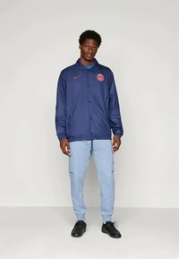 Vīrietis, kurš valkā tumši zilu pogājamu žaketi ar sarkaniem logotipiem, gaiši zilas cargo bikses un melnas sporta kurpes, stāv pretī vienkāršai gaišai fona.