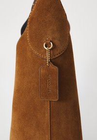 Bruine suède handtas met een ronde klep en een gouden kettinglabel, met "COACH NEW YORK" erop geprezen, tegen een effen achtergrond.