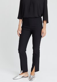 Frau in schwarzen, figurbetonten Hosen mit vorderen Knöchelschlitzen und silbernen Slingback-Ballerinas vor einem schlichten hellen Hintergrund.