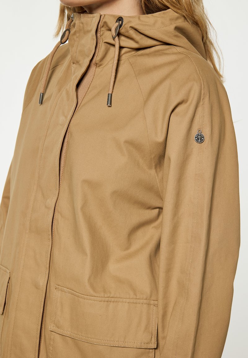 Veste kaki avec capuche, dotée d'une fermeture à cordon de serrage et de poches latérales. Texture lisse et design minimaliste avec un petit détail en métal au logo.