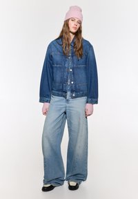 Giubbotto di jeans con chiusura a bottoni, jeans blu a gamba larga e berretto in maglia rosa. Il giubbotto presenta un taglio classico e tasche frontali.