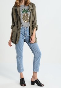 Chaqueta militar de color verde oliva sobre una camiseta gris con un diseño de piña de lentejuelas; jeans de pierna recta de color azul claro y mule de tacón bloque negro.