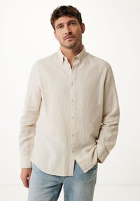 Chemise boutonnée beige clair en lin, dotée d'un col traditionnel, de manches longues et d'un ourlet légèrement arrondi, associée à un jean bleu clair.