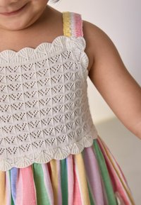 Robe tricotée avec un bustier texturé blanc présentant une bordure en zigzag ; jupe superposée à rayures multicolores dans des teintes pastel de rose, vert et jaune.