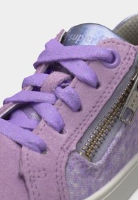 Gros plan d'une basket violette avec des lacets texturés, un matériau semblable au daim, une fermeture éclair en métal et un détail métallique au niveau du col avec le mot « superfit ».