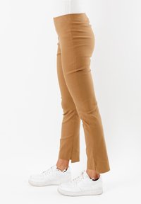 Pantaloni marroni su misura realizzati in tessuto elasticizzato, caratterizzati da una vestibilità slim e da spacchi laterali all'orlo. Abbinati a sneakers bianche.