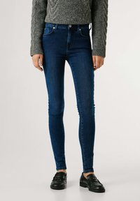 Vaqueros ajustados azul oscuro hechos de mezclilla con un ajuste elegante, que cuentan con cinco bolsillos y cierre de botón, usados con mocasines negros.