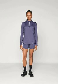 Kvinde står iført en lilla langærmet zip-up bluse, matchende shorts, sorte sokker og sorte sportsko foran en ensfarvet baggrund.