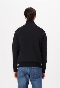 Sweatshirt noir avec un col montant, poignets et bord côtelés, fabriqué en tissu lisse ; porté avec un jean bleu avec poches arrière.
