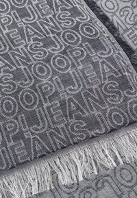 Denimový šál s opakovaným textom značky "JOOP! JEANS" v svetlo šedej farbe na tmavom pozadí, s strapcami na koncoch.