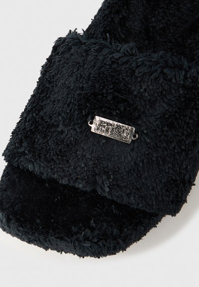 Juoda tekstūruota fliso pirštinė su mažu stačiakampiu metalo ženkleliu, kuriame yra skaičiai ir apskritimu pažymėtas skaičius šeši riešo srityje.