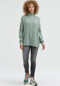 Hellgrüner Strickpullover mit Rollkragen und Rippenbündchen, kombiniert mit dunkelgrauen Skinny-Jeans und türkisfarbenen Turnschuhen mit pinken Akzenten.