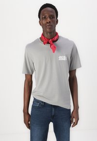 Graues Baumwoll-T-Shirt mit kurzen Ärmeln, das ein weißes Logo auf der Brust trägt. Kombiniert mit dunkelblauen Jeans und einem roten Halstuch um den Hals.