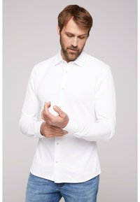 Camicia bianca a maniche lunghe con colletto abbottonato e patta frontale, abbinata a jeans azzurri; tessuto morbido e design aderente.
