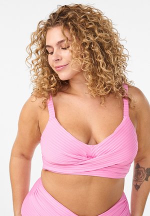 Krøllet kvinde med lyserød ribstrikket crop top og matchende bund, der smiler blidt, med en rosetatovering på venstre arm.