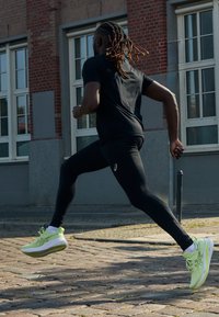 Maglietta atletica nera e leggings aderenti indossati mentre si corre su ciottoli. Scarpe da corsa verde chiaro con accenti bianchi e suole texture.