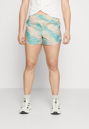 Kvinde iført beige og turkismønstrede sportsshorts, hvide sokker og hvide sneakers, stående mod en ensfarvet lys baggrund.