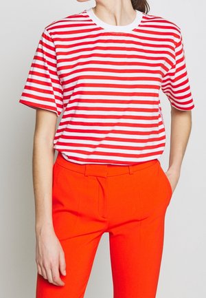 Persoon die een rood-wit gestreept shirt met korte mouwen en felrode broek draagt, met één hand in de zak en een neutrale achtergrond.