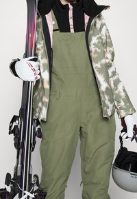 Skiausrüstung mit einem hellolivgrünen Overall, einer jacke mit floralen Mustern, weißen Handschuhen mit roten Akzenten und Skiausrüstung in der Hand.
