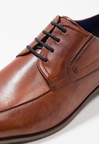 Chaussure en cuir marron au fini lisse, dotée de lacets noirs et de subtils détails de couture. Bout arrondi et design à profil bas.