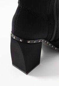 Schwarze Knöchelstiefel aus dehnbarem Material mit blockigem Absatz. Mit einem genoppten schwarzen Riemen um den Knöchel und einem seitlichen Reißverschluss.