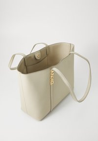 Borsa a tracolla beige con superficie liscia, due manici lunghi e un accento con logo dorato. Presenta una tasca interna e una chiusura a bottone.