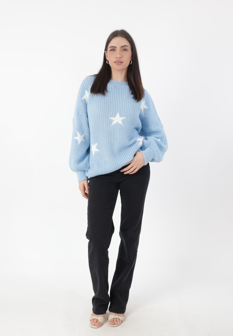 Lichtblauwe gebreide trui met witte sterpatronen, brede mouwen en een ronde halslijn, gecombineerd met zwarte broek en lichte sandalen.