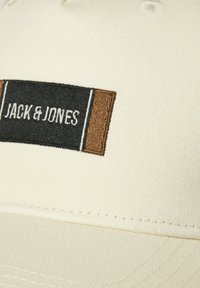 Close-up van een beige stof met een zwart, bruin en wit geborduurd label met de tekst "JACK & JONES" erop gestikt.