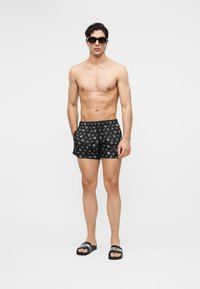 Skjorteløs mann iført svarte badeshorts med mønster, svarte solbriller og svarte sandaler med hvit tekst på en ensfarget hvit bakgrunn.
