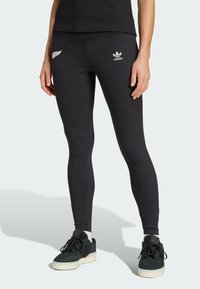 Leggings neri realizzati in tessuto elastico con un logo bianco a forma di felce e branding Adidas, abbinati a sneakers bianche e nere.