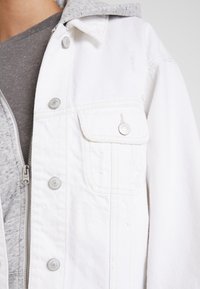 Chaqueta de mezclilla blanca con cierres de botones y un bolsillo en el pecho, superpuesta sobre una sudadera con capucha gris, con un tejido texturizado.