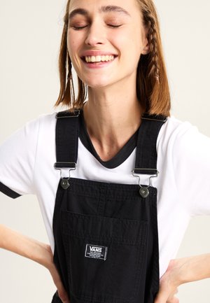 Sorte bomulds overalls med metal clips, med en frontlomme og et Vans logo patch. Båret over en hvid t-shirt med sort kant.