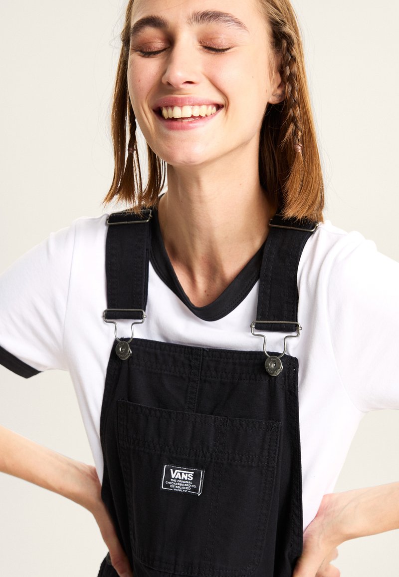 Salopette en coton noire avec des clips en métal, avec une poche avant et un écusson logo Vans. Portée par-dessus un t-shirt blanc avec des bordures noires.