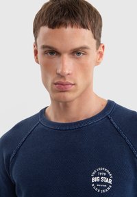 Námořnická modrá mikina s kulatým výstřihem, vyrobená z texturovaného pleteného materiálu, s raglánovými rukávy a kruhovým bílým logem na hrudi.