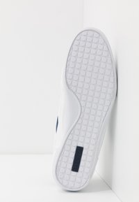 Baskets blanches avec une semelle en caoutchouc texturé présentant des motifs circulaires, un dessus en cuir lisse, et un accent sombre avec un logo.