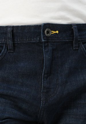 Jeans in denim blu scuro con chiusura a bottone standard con un accento giallo e multiple tasche. La texture appare liscia con una leggera lucentezza.