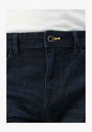 Tmavě modré džíny z denimu s klasickým zapínáním na knoflík se žlutým akcentem a několika kapsami. Povrch vypadá hladce s mírným leskem.