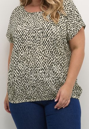 Kvinde iført en beige kortærmet top med sorte uregelmæssige pletter og mørkeblå jeans, stående mod en ensfarvet lys baggrund.