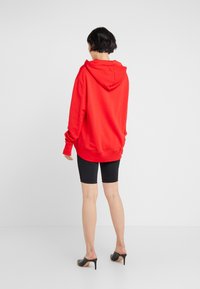 Punainen huppari leveällä hupulla ja resorilla varustetuilla hihoilla, yhdistettynä mustiin pyöräilyshortseihin. Kangas näyttää pehmeältä ja istuu rentona. Kengät ovat mustat korkokengät.