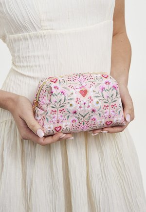 Ruke drže ružičastu torbicu s uzorkom cvijeća i srca, preko bijele teksturirane haljine.