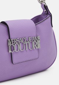 Fioletowa skórzana torebka z teksturowaną powierzchnią, z metalowym logotypem "VERSACE JEANS COUTURE" oraz usztywnionym zapięciem na klapkę.