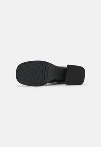Semelle de chaussure à talon noire avec motif ondulé et un talon large et carré sur fond blanc.
