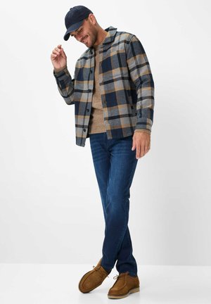 Kariertes Flanellhemd in Blau, Grau und Braun, über einem beigen Pullover getragen, kombiniert mit blauen Jeans und beigefarbenen Schuhen, ergänzt durch eine marineblaue Kappe.