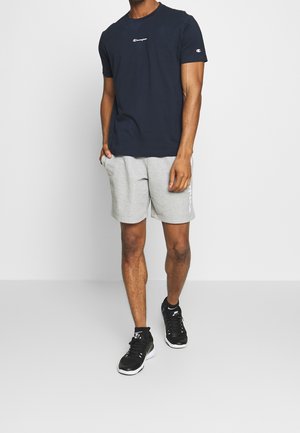 Mann trägt ein marineblaues Champion-T-Shirt, hellgraue Champion-Shorts und schwarze Sportschuhe, steht vor einem schlichten weißen Hintergrund.