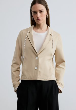 Woman wearing beige buttoned jacket with drawstrings over white top and black pants, standing with hands in pockets against plain background.