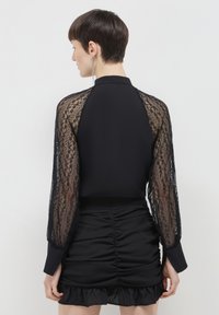 Frau mit kurzen Haaren, die ein schwarzes Kleid mit durchsichtigen, langen Spitzenärmeln und gerafftem Rock trägt, von hinten vor weißem Hintergrund gezeigt.