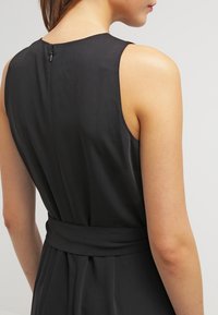 Svart ärmlös klänning med en åtsittande överdel, bälte i midjan och dold dragkedja bak. Slät, matt textur och en blygsam silhuett.