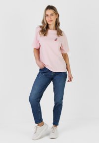 Tânără femeie stând cu mâna în buzunar, purtând un tricou roz deschis cu un model de cireșe, blugi albaștri și tenisi albi, pe un fundal simplu.